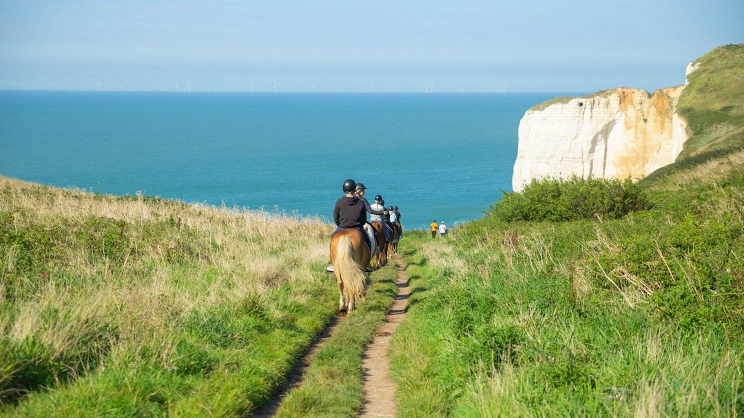 a group of riders pictured in single file from behind, descending a coastal path.

There is blue sea and a white cliff protruding into it in the distance